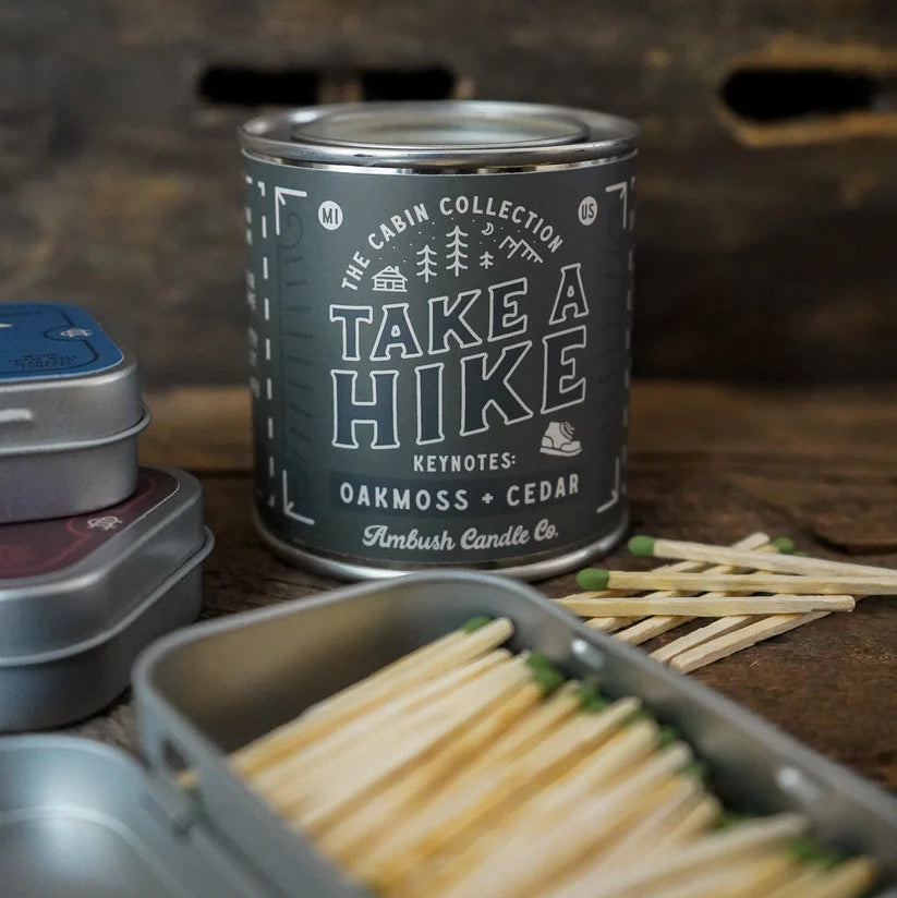 A metal tin of matches labeled 'Take a Hike' by Ambush Candle Co., with wooden matchsticks and additional metal tins on a rustic dark wood surface.