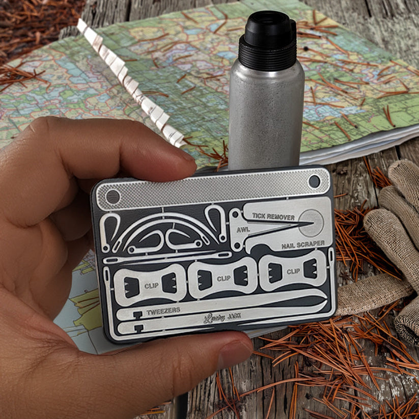 A hand holds a silver and black multi-tool card case over a rustic wooden table with pine needles, a map, gloves, and a metal bottle.