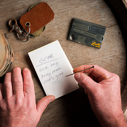 A top-down view of a person's hands writing on a small white notepad placed on a rustic wooden barrel. A vintage lantern, a leather key fob, and a cassette tape are nearby on the wood.